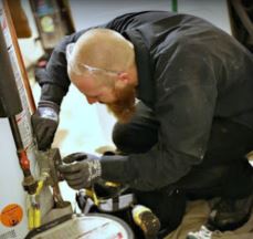 Plumber wearing safety glasses and gloves, crouched down and working on a water heater, with tools and a vacuum nearby.